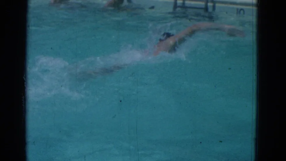1953 Yonkers pool footage shows the swimmer mid-stroke, with a pool ladder and edge markings faintly visible above the water.