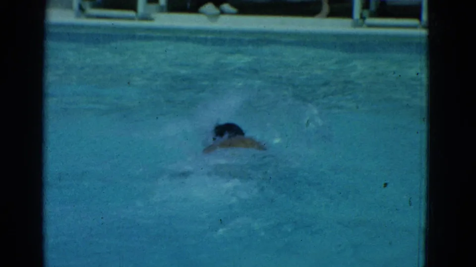 A swimmer moves toward the camera in blue pool water, with railing and faint figures beyond the pool.