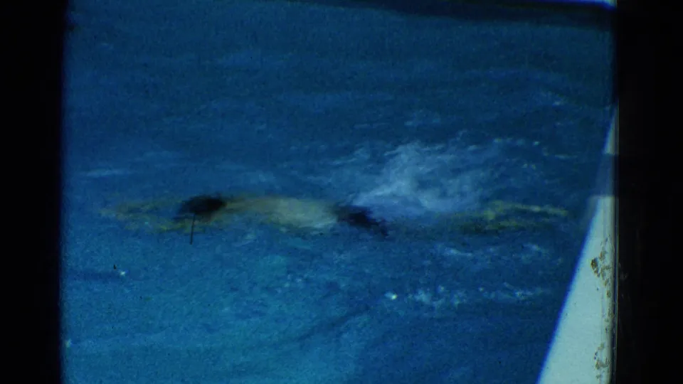 A swimmer in lighter swimwear moves through darker blue water near the pool edge in the 1953 footage.