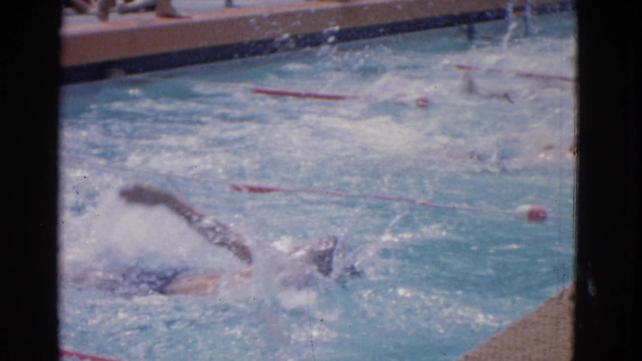 Backstroke swimmers move through a pool beside red lane dividers and a textured deck.