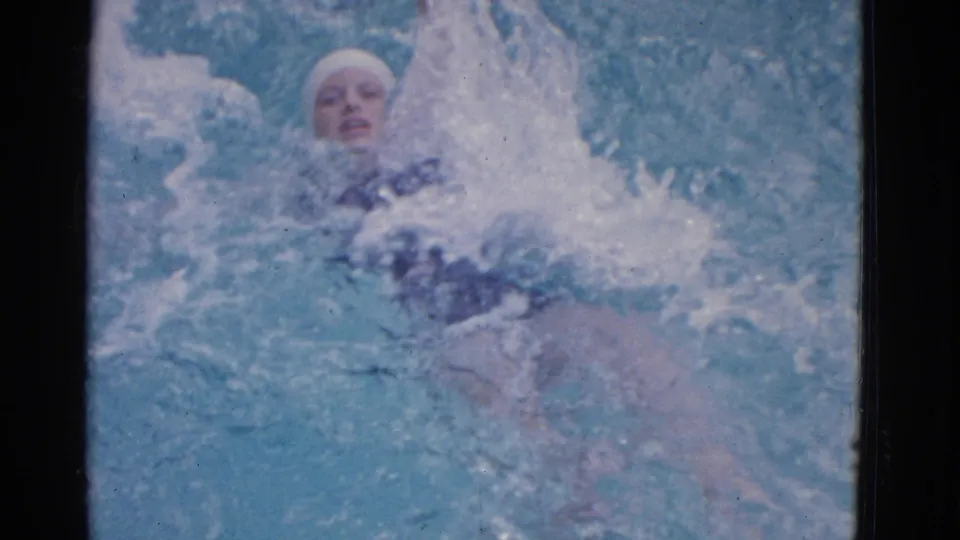 1953 Yonkers frame showing the woman swimmer's face, white swim cap, and dark swimsuit amid splashing blue pool water.