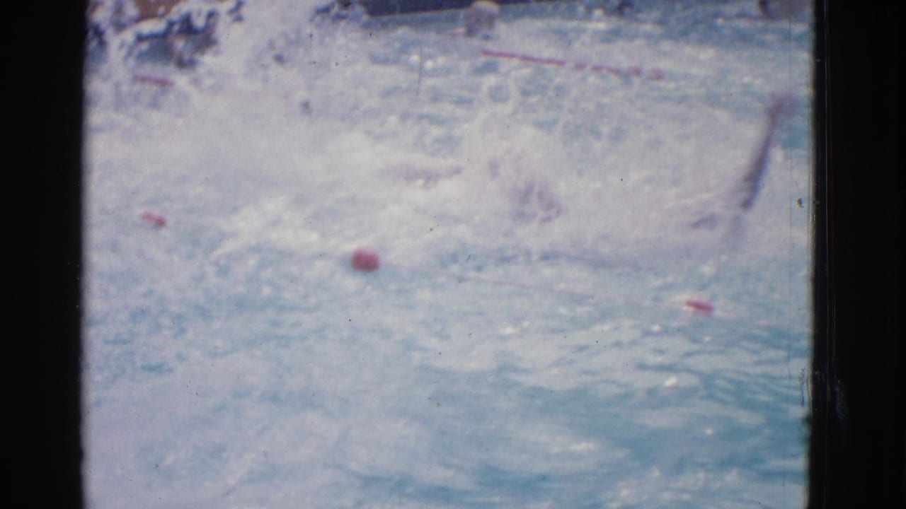 Swimmer partly submerged in a pool lane with splashing water and red buoys.