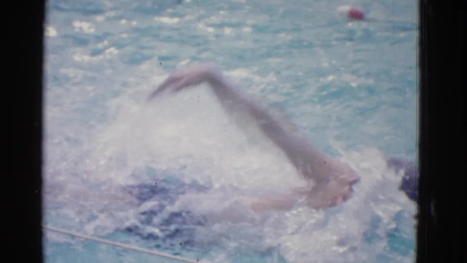 A young swimmer continues backstroke practice in the pool, with his arm extended overhead and his body partly obscured by spray.