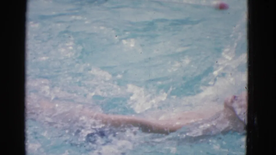 The child swimmer appears stretched across the lower pool frame during backstroke practice, with water spray and a dark window divider nearby.
