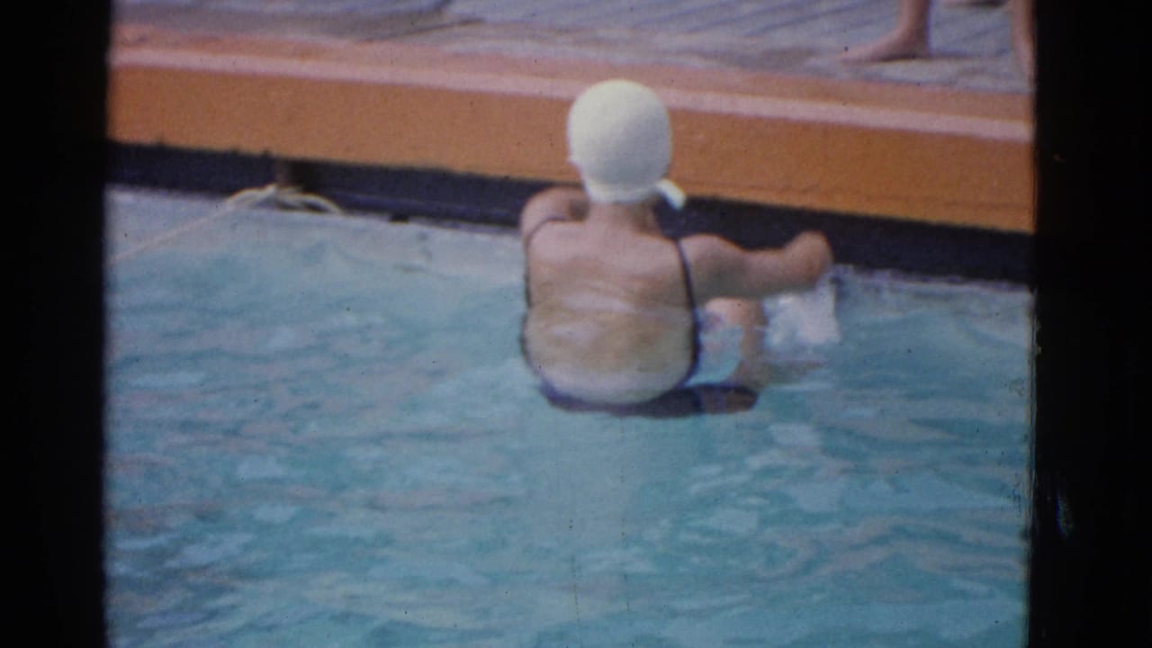 1953 Yonkers home-movie frame showing a swimmer in a white cap and dark swimsuit near the edge of a turquoise backyard pool.