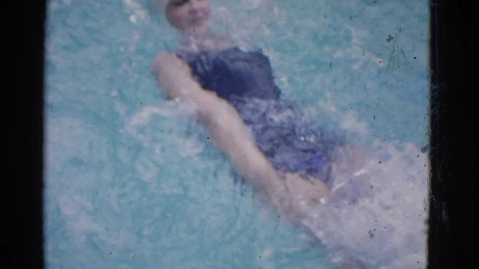 1953 Yonkers home-movie frame showing a swimmer in a dark suit and white cap turning toward the camera in the pool.