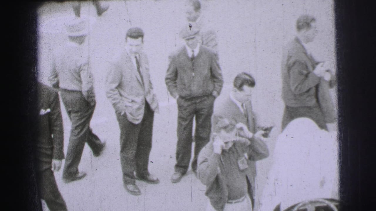 At Le Mans in 1954, adult men in suits, jackets and caps stand near race cars during track-side preparation.