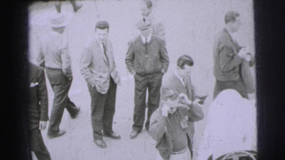 At Le Mans in 1954, adult men in suits, jackets and caps stand near race cars during track-side preparation.