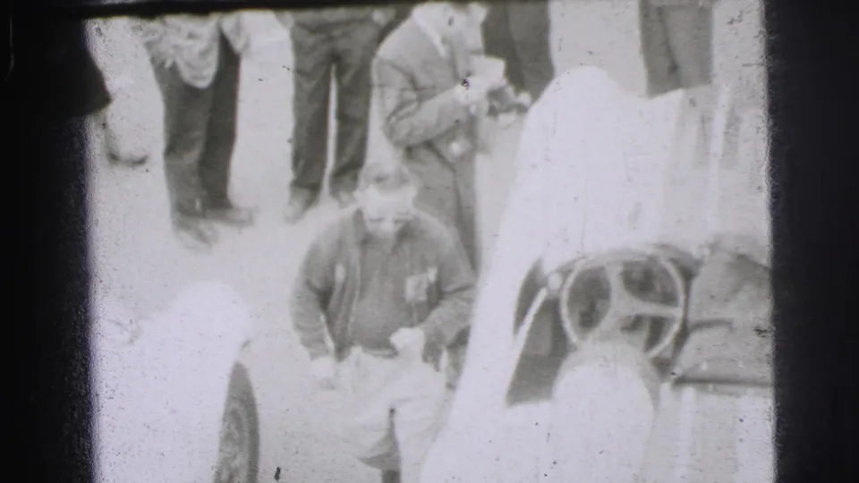 At the Le Mans track in 1954, adult men gather around an open car while a man in a sweater stands in the foreground.
