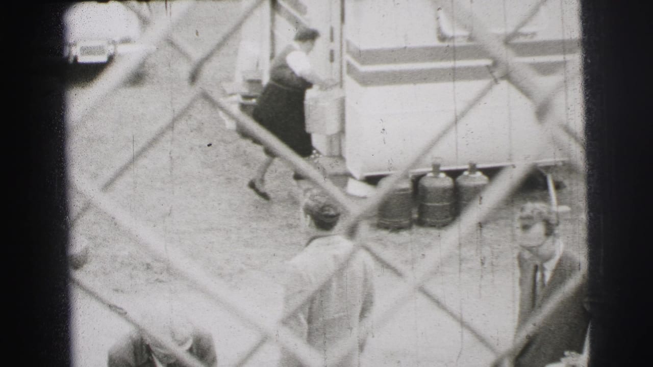 Workers and trailers seen through a chain-link fence, with small cylinders near the trailer.