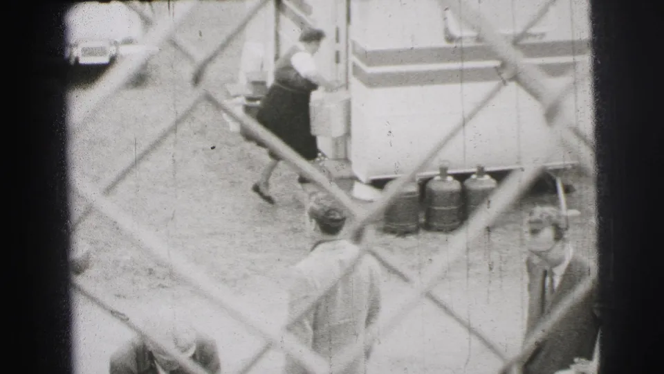 Le Mans, France, 1954 home-movie frame showing workers near trailers and supplies behind a chain-link fence.