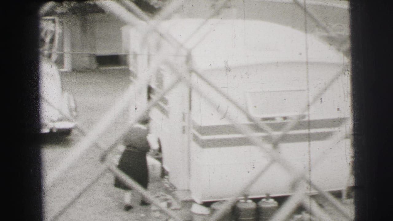 Le Mans, France, 1954 frame with workers near a large trailer, cylinders, and a partly visible vehicle beyond the fence.