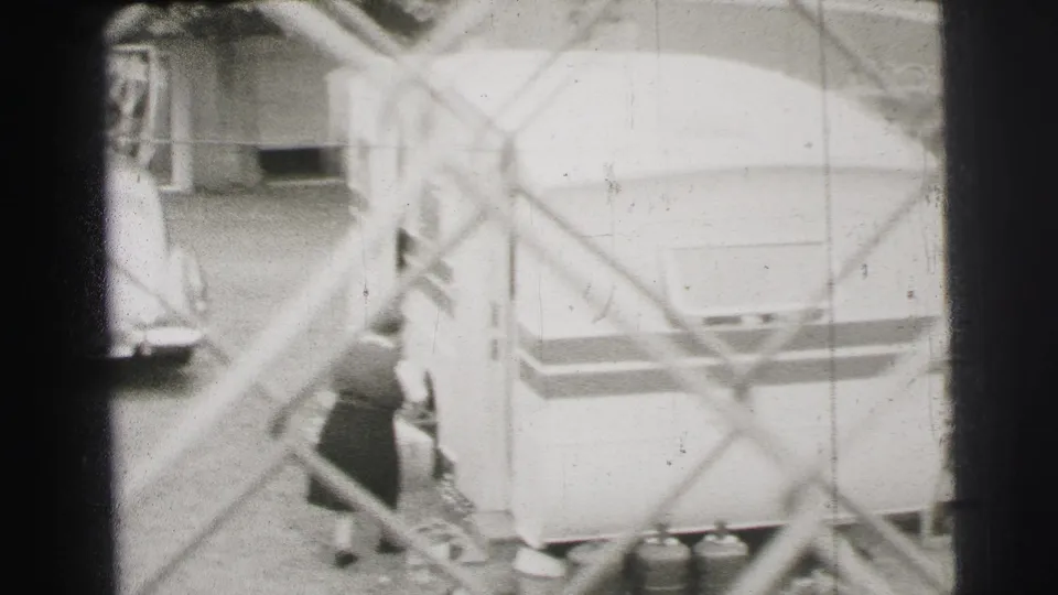 Le Mans, France, 1954 frame with workers near a large trailer, cylinders, and a partly visible vehicle beyond the fence.