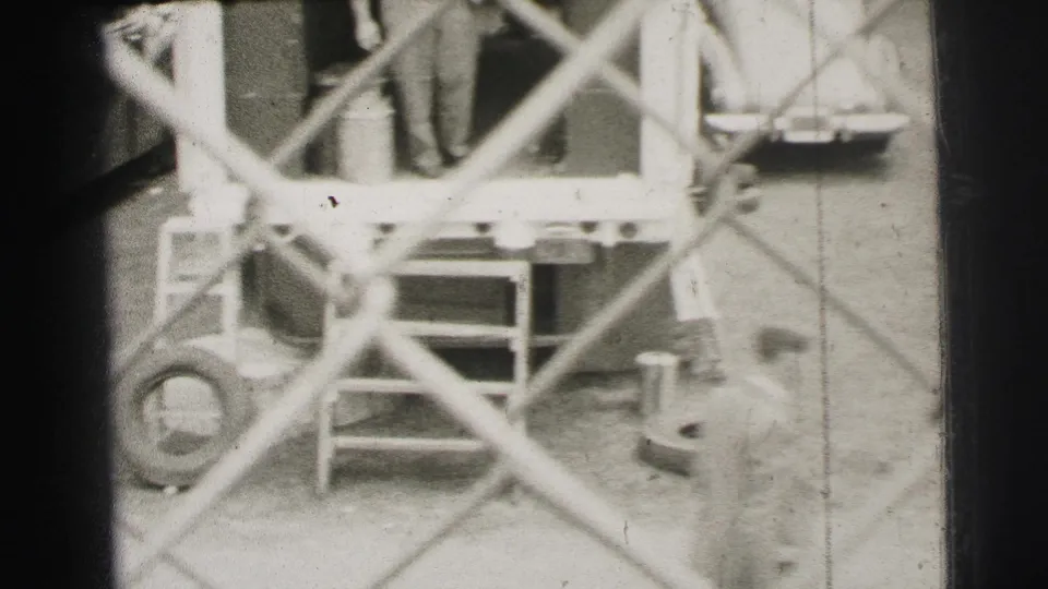 Le Mans, France, 1954 frame showing trailer steps, workers, hoses or cables, and site equipment through chain-link fencing.