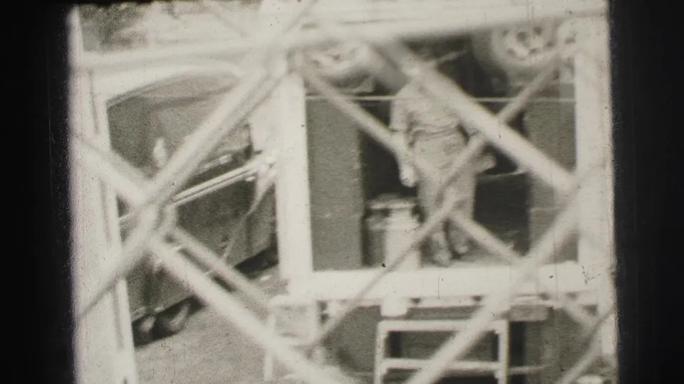 Le Mans, France, 1954 frame showing a worker near a trailer opening, vehicle parts, and equipment behind chain-link fencing.
