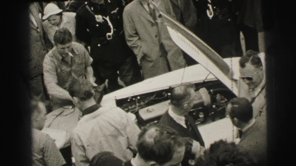 At Le Mans in 1954, men in suits and casual clothes crowd around a race car with its hood open and engine area exposed.