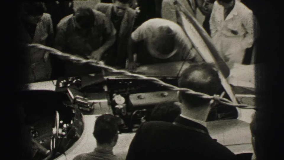 At the 1954 Le Mans event, a mechanic appears to work over the car as onlookers stand tightly around the open hood.