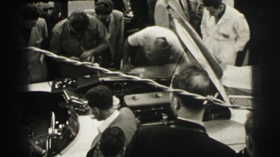 The 1954 Le Mans footage shows a mechanic leaning over the race car while spectators and officials cluster around the open hood.
