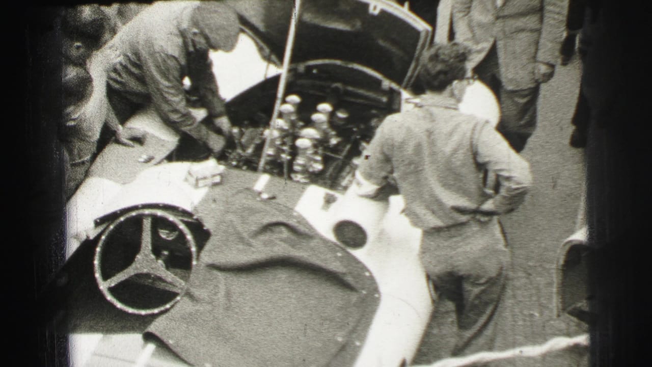 Mechanics lean over the open engine bay of a light race car as spectators stand nearby.