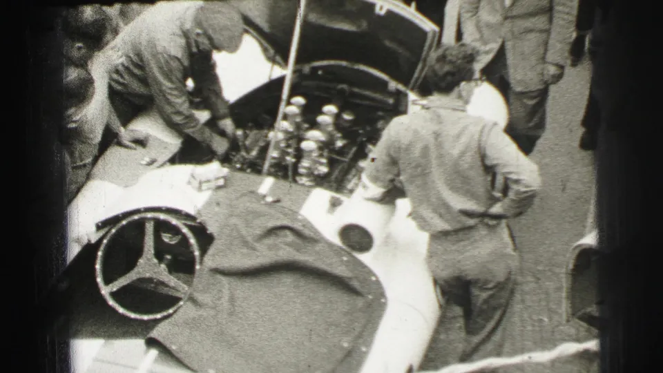 At Le Mans in 1954, mechanics work around the open hood of a light Mercedes-Benz race car while spectators watch close by.