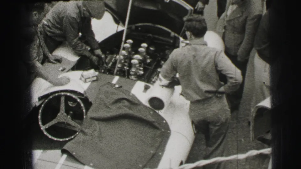 At the 1954 Le Mans setting, a mechanic reaches into the open engine area of a Mercedes-Benz race car as onlookers gather.
