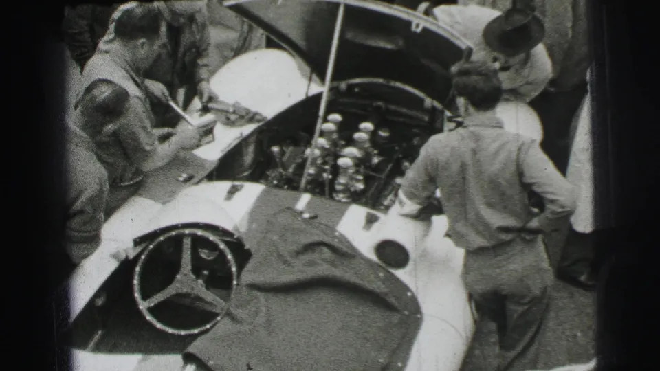 Several men cluster around the open engine bay of a light Mercedes-Benz race car during the 1954 Le Mans footage.