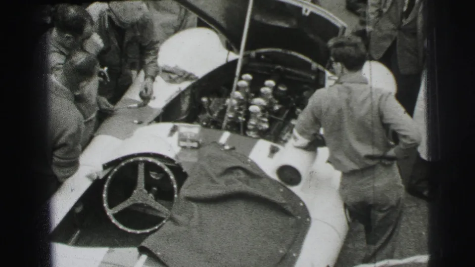 At Le Mans in 1954, mechanics work under the hood of a Mercedes-Benz race car as spectators and crew gather close by.