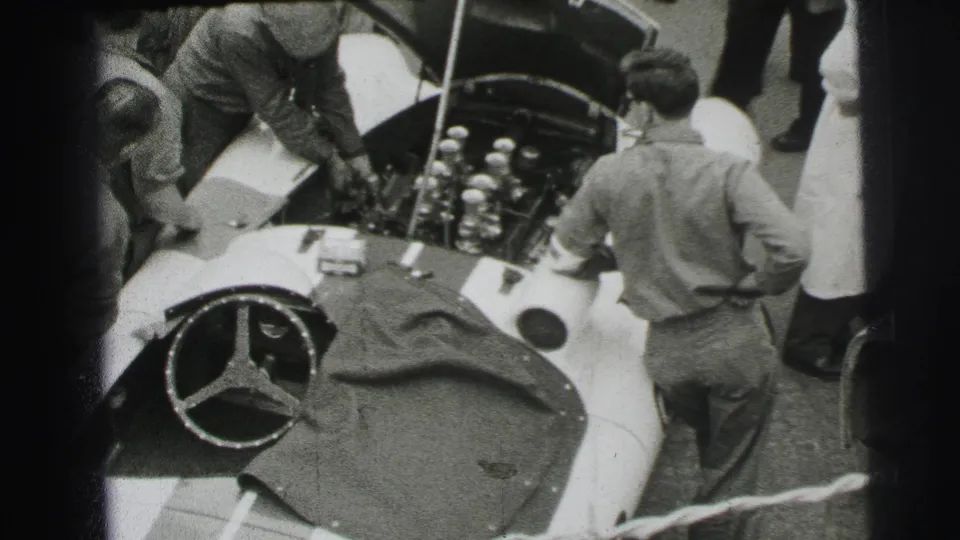 A 1954 Le Mans pit-lane view shows crew members leaning into a Mercedes-Benz race car with its hood raised.