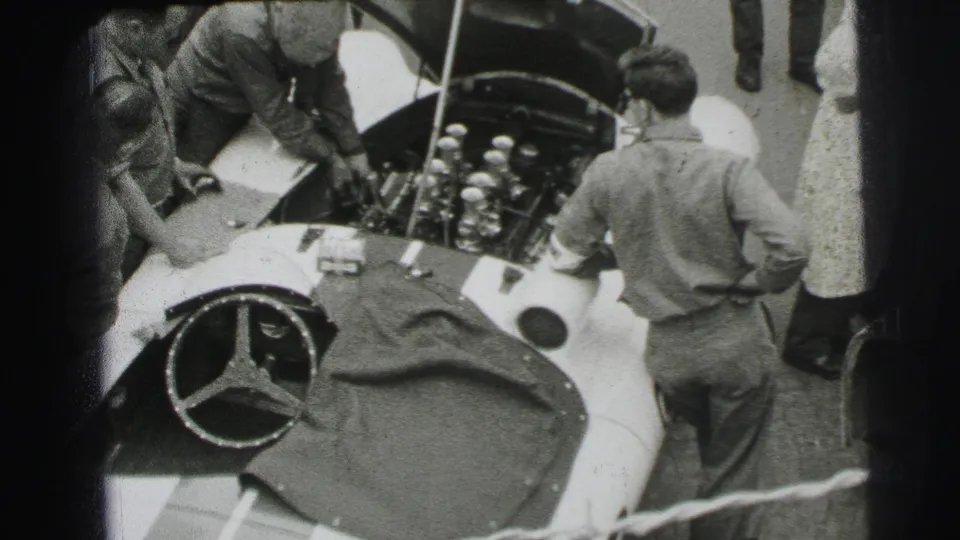 At the Le Mans pit lane, men gather around a Mercedes-Benz race car while mechanics inspect the open engine compartment.
