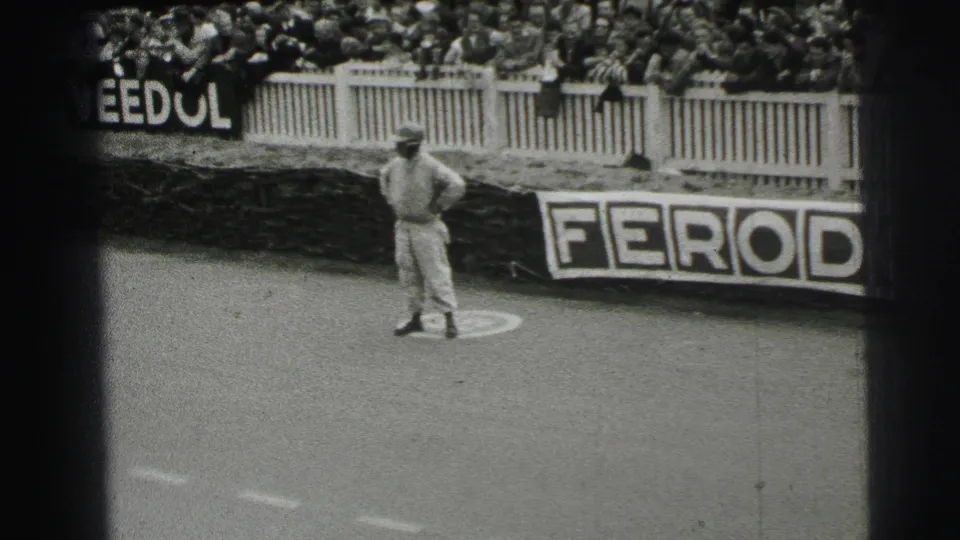 A 1954 Le Mans trackside view shows a man in light clothing standing before crowded barriers and visible advertising boards.