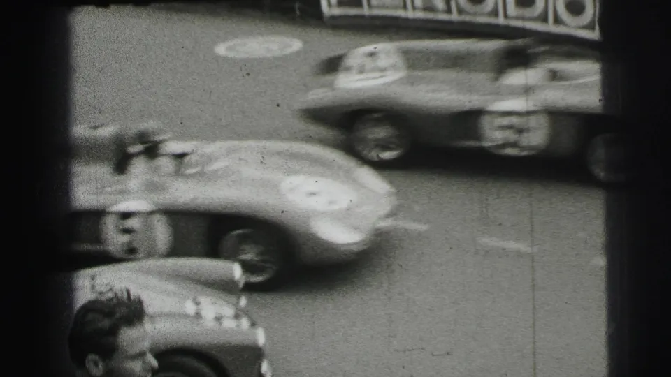 Black-and-white 1954 Le Mans home-movie frame showing blurred racing cars near Veedol trackside advertising and a spectator edge.