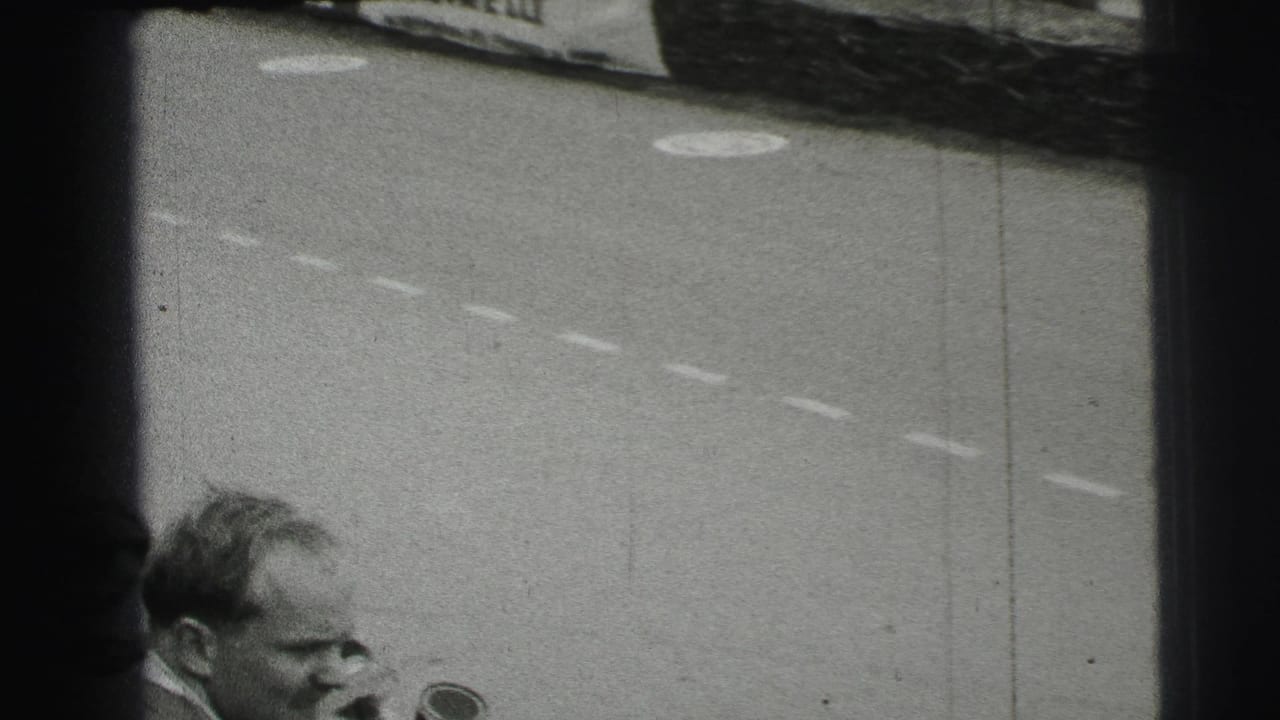 Black-and-white 1954 Le Mans frame with a nearby male spectator or trackside figure in front of the circuit and a partial racing car.