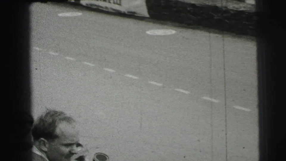 Black-and-white 1954 Le Mans frame with a nearby male spectator or trackside figure in front of the circuit and a partial racing car.