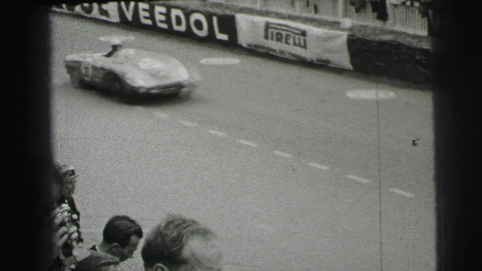 Black-and-white 1954 Le Mans frame showing a low racing car on the track beside Veedol and Pirelli advertising boards.