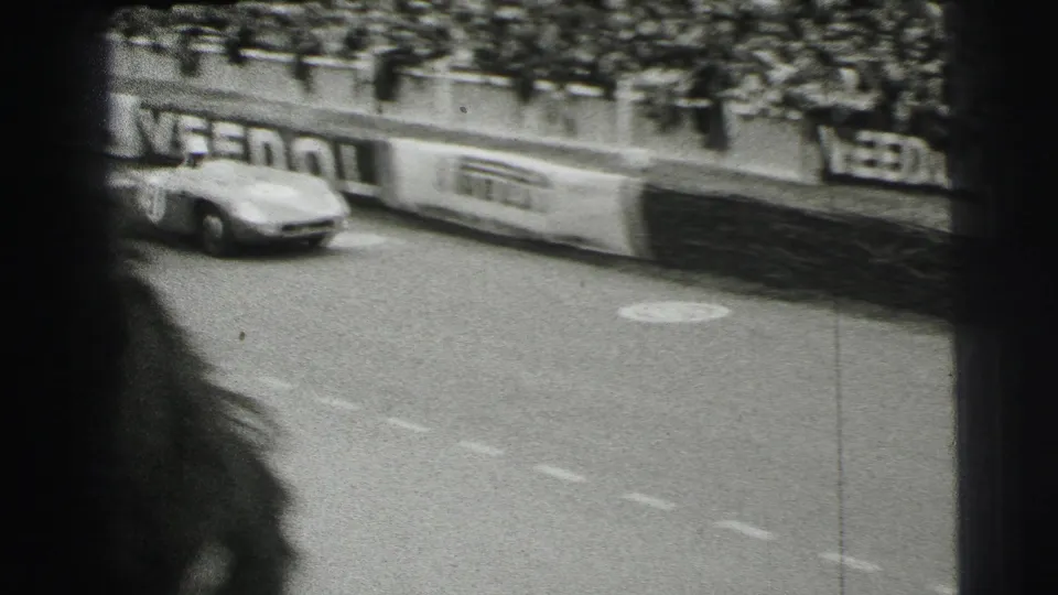 Black-and-white 1954 Le Mans home-movie frame showing a racing car below a dense spectator crowd and Veedol trackside signage.