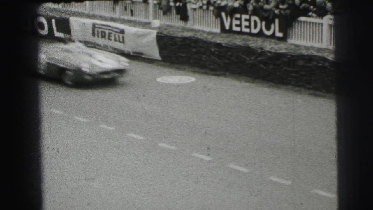 A single sports car passes quickly on the Le Mans track, with crowd barriers and period advertising visible in the background.