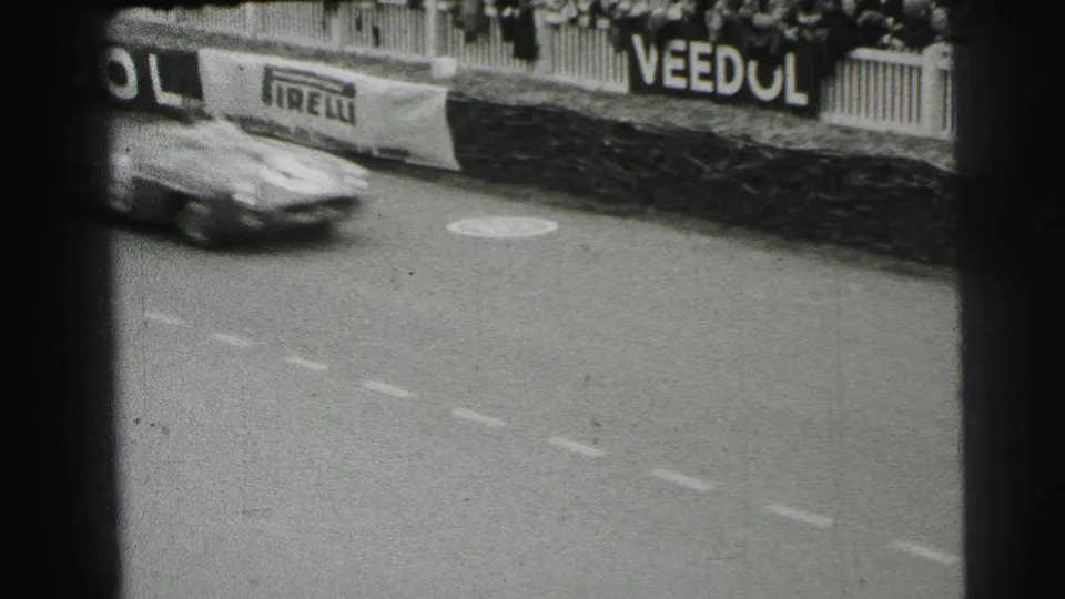 A single sports car passes quickly on the Le Mans track, with crowd barriers and period advertising visible in the background.