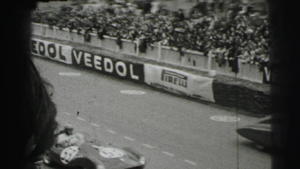 A spectator viewpoint over the Le Mans course shows crowded barriers, Veedol and Pirelli signage, and a race car partly visible at the bottom.