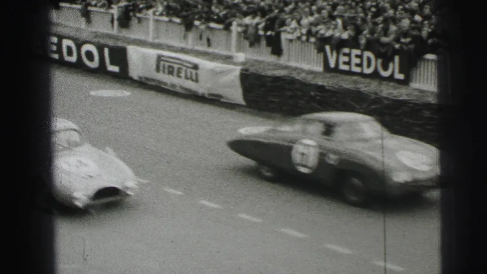 At Le Mans in 1954, blurred race cars speed past packed stands and trackside advertising barriers on black-and-white 8mm film.