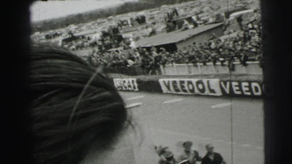 Spectator-shot 1954 Le Mans footage with a foreground head or hat obscuring part of the stands and branded barriers.