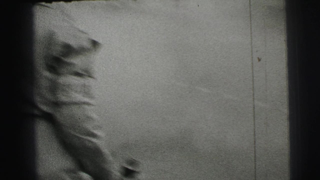 A cropped spectator or passerby appears close to the camera, with the racetrack surface and film scratches visible in the 8mm frame.