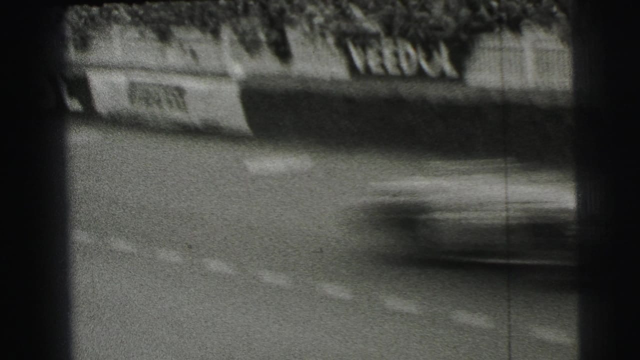 A blurred race car speeds past Le Mans trackside sponsor boards, with fencing and spectators visible in the background.