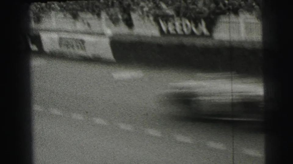A blurred race car speeds past Le Mans trackside sponsor boards, with fencing and spectators visible in the background.