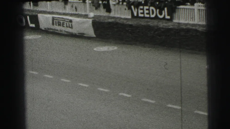 Le Mans trackside view after a car has passed, showing sponsor boards, barrier fencing, road markings, and grainy 8mm texture.