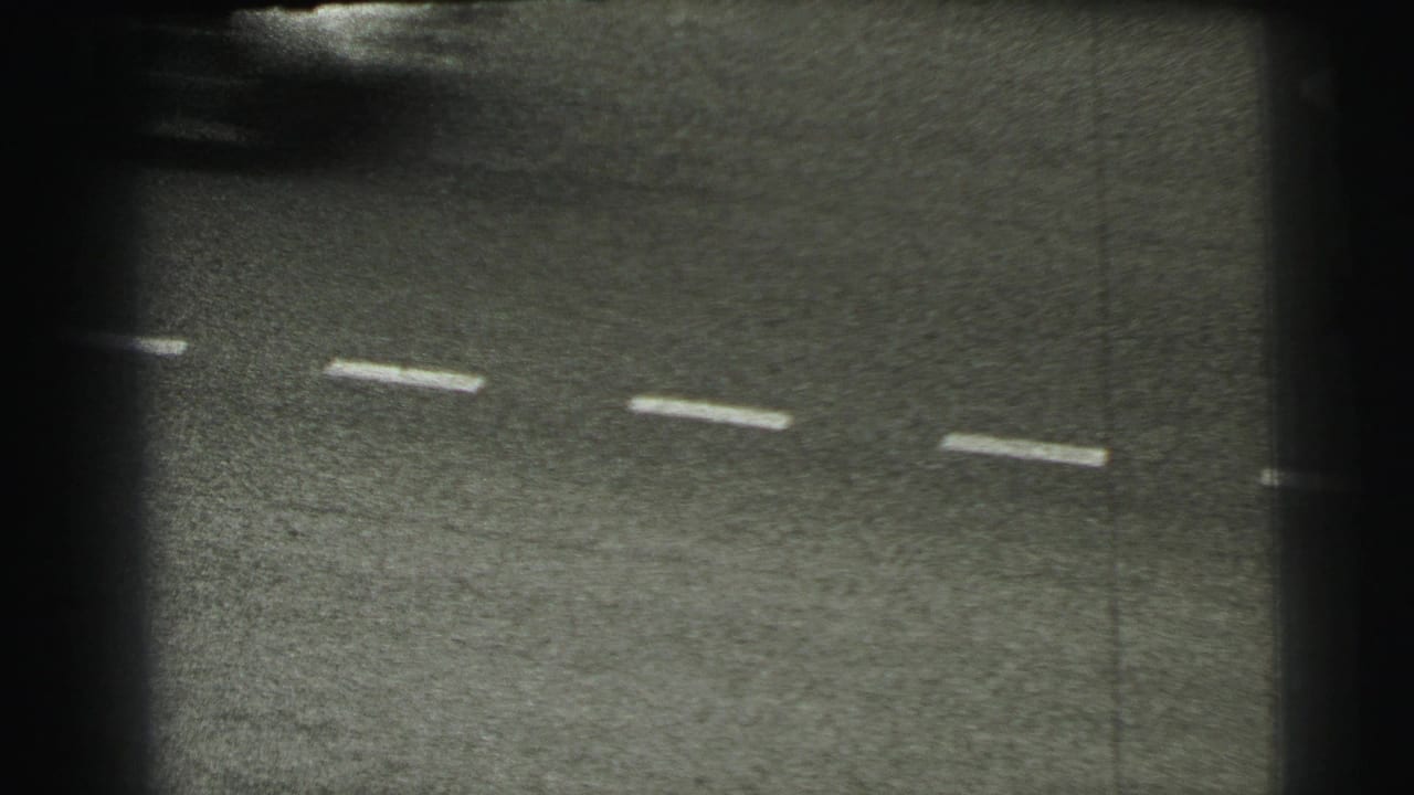 Grainy 1954 Le Mans frame focused on the asphalt and dashed markings, with heavy vignette and partial trackside signage at the edge.