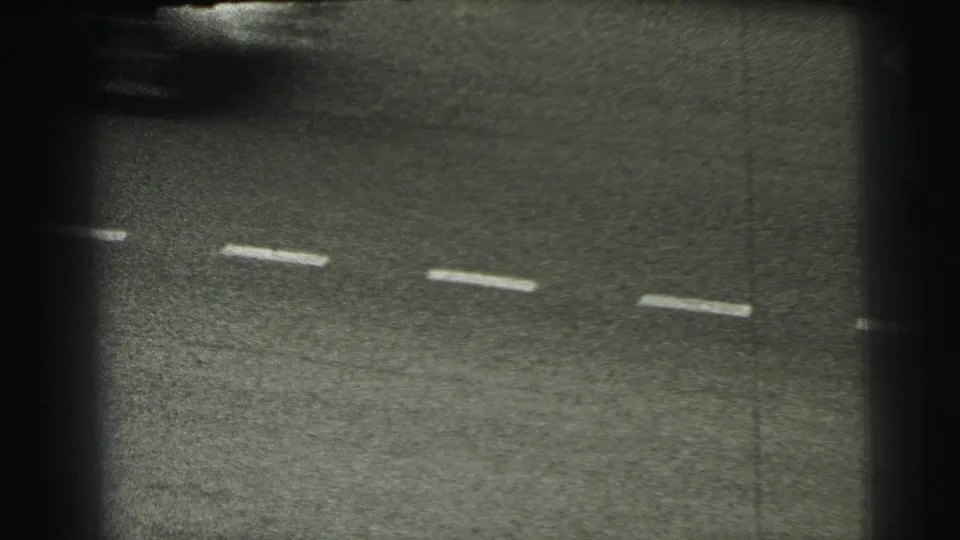 Grainy 1954 Le Mans frame focused on the asphalt and dashed markings, with heavy vignette and partial trackside signage at the edge.