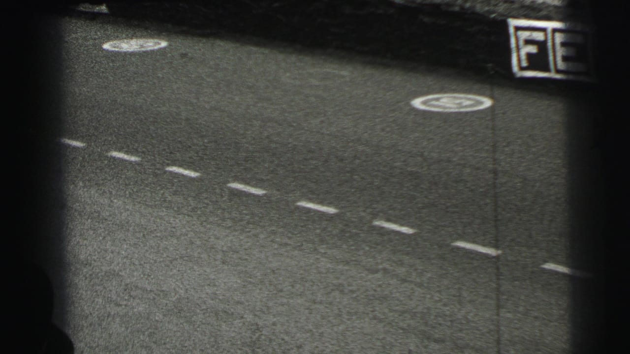 1954 Le Mans 8mm frame with empty marked asphalt, circular painted symbols, and a cropped trackside advertising board on the right.