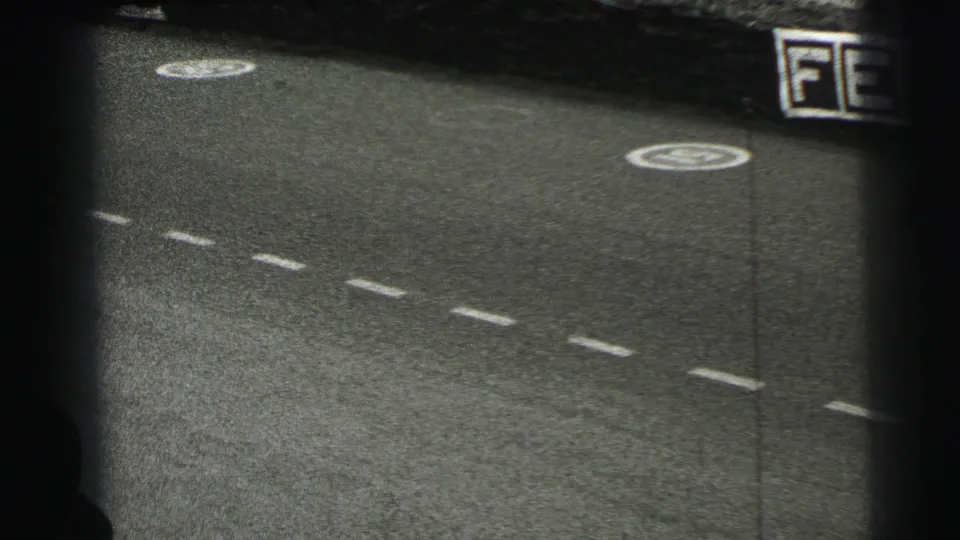1954 Le Mans 8mm frame with empty marked asphalt, circular painted symbols, and a cropped trackside advertising board on the right.