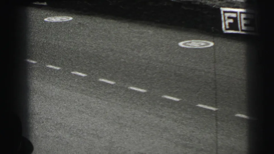 1954 Le Mans home-movie frame showing a paved track, dashed white markings, circular painted symbols and partial lettering near the edge.