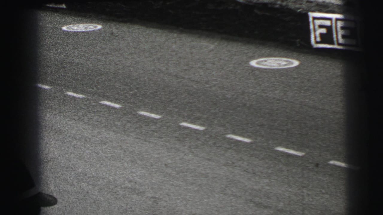 1954 Le Mans frame with the race surface, dashed lane markings, circular symbols and a dark blurred passing vehicle at the edge.